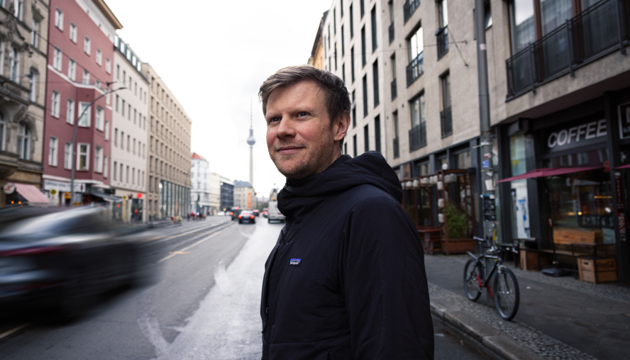 Unser Kollege Bastian Pfahl steht auf einer Straße in Berlin. In der Flucht erkennt man den Berliner Fernsehturm. Er lächelt freundlich in die Kamera. 