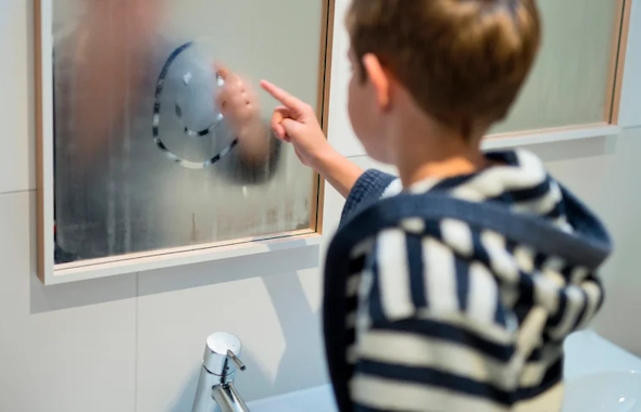 Ein Kind im gestreiften Oberteil malt einen Smiley an einen beschlagenen Spiegel im Bad. 
