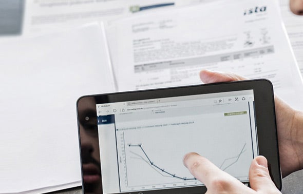 Eine Männerhand zeigt auf ein Tablet auf dem ein Graph zu erkennen ist. Im Hintergrund sind leicht verschwommen Dokumente der ista erkennbar. 
