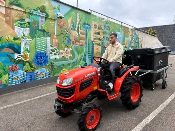 Ein Mann fährt mit einem roten Minischlepper samt Anhänger vor einer bunt bemalten Wand mit Naturmotiven.