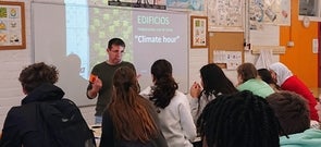 Ein Lehrer erklärt einer Gruppe von Schüler:innen im Klassenzimmer das Projekt „Climate hour“ zur klimafreundlichen Gebäudenutzung; im Hintergrund ist eine Präsentation mit dem Titel „EDIFICIOS respetuosos con el clima“ zu sehen.