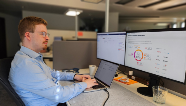 Ein Mann mit hellbraunem Haar und Brille sitzt an einem Schreibtisch in einem modernen Büro und arbeitet an einem Laptop. Auf einem der zwei Monitore vor ihm ist ein Diagramm zu sehen. Auf dem Schreibtisch befinden sich eine Tasse und ein Glas.