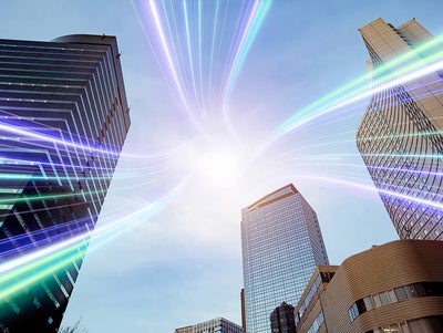 The image showcases a dynamic urban scene featuring several modern skyscrapers reaching towards the sky. The buildings are a mix of glass and steel structures, reflecting a professional and corporate environment. Superimposed over the scene are vibrant, multicolored light streaks that converge at a bright point in the sky, symbolizing connectivity and technology. The light streaks add a futuristic and energetic element to the image, suggesting advanced communication networks or data transfer within a bustling cityscape. The overall composition highlights the intersection of modern architecture and innovative technology.