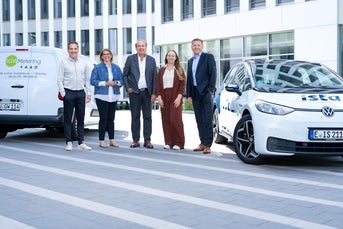 Fünf Personen stehen lächelnd nebeneinander vor einem modernen Bürogebäude. Links ist ein weißer Transporter mit der Aufschrift „SGW Metering“ zu sehen, rechts ein weiß-blaues Elektroauto von „ista“. Die Gruppe wirkt geschäftlich gekleidet und symbolisiert eine Kooperation zwischen den beiden Unternehmen.