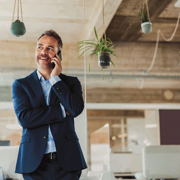 The image depicts a professional setting, likely an office. A man in a dark blue suit, with a light blue shirt underneath, is standing and talking on his mobile phone. He appears to be in a modern, open-concept office space with natural light coming in, creating a bright and airy environment. The office has an industrial design with exposed concrete ceilings and hanging plants adding a touch of greenery. The man looks content and engaged in his conversation, with a slight smile on his face. In the background, there are desks, chairs, and computer monitors, indicating a busy work atmosphere. The overall scene suggests a modern, dynamic workplace.