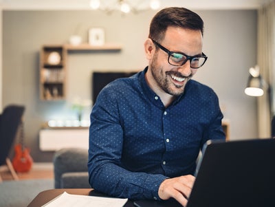The image depicts a man sitting at a round table in a well-lit office space. There is a glass of water and a small espresso cup on the table, adding to the professional yet comfortable atmosphere. The man is smiling and engaged with the water consumption billing from ista on his laptop, taking notes in a notebook. He is wearing a blue button-down shirt and glasses. The background features a flip chart, potted plants, modern furniture, and large windows allowing natural light to flood the room. 