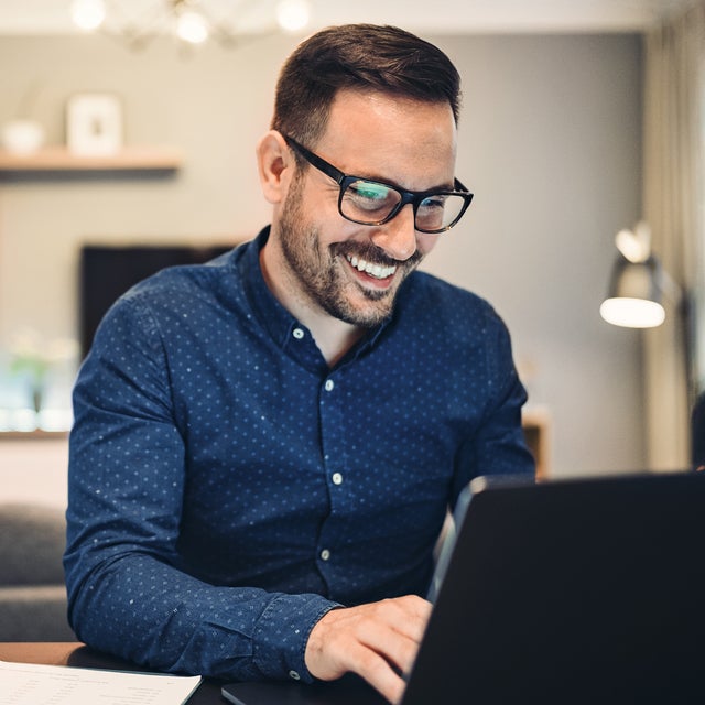 The image depicts a man sitting at a round table in a well-lit office space. There is a glass of water and a small espresso cup on the table, adding to the professional yet comfortable atmosphere. The man is smiling and engaged with the water consumption billing from ista on his laptop, taking notes in a notebook. He is wearing a blue button-down shirt and glasses. The background features a flip chart, potted plants, modern furniture, and large windows allowing natural light to flood the room. 