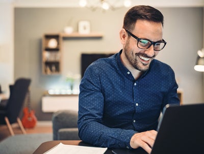 The image depicts a man sitting at a round table in a well-lit office space. There is a glass of water and a small espresso cup on the table, adding to the professional yet comfortable atmosphere. The man is smiling and engaged with the water consumption billing from ista on his laptop, taking notes in a notebook. He is wearing a blue button-down shirt and glasses. The background features a flip chart, potted plants, modern furniture, and large windows allowing natural light to flood the room. 