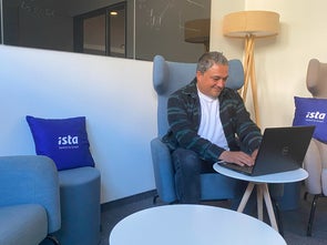 Ein Mann arbeitet konzentriert an einem Laptop in einer gemütlichen Sitzecke mit blauen Sesseln und Kissen mit dem Aufdruck „ista – Switch to Smart“. Im Hintergrund ist eine Stehlampe und eine Tafel mit Zeichnungen zu sehen.