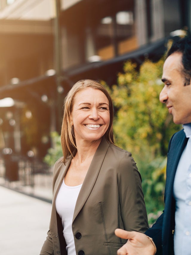 Two business professionals, a man and a woman, engage in a friendly conversation while walking outside near modern office buildings. The woman, dressed in a smart blazer, smiles warmly, while the man gestures as he speaks. The background features greenery and urban architecture, suggesting a productive, collaborative environment in a professional setting. The image reflects themes of business communication, networking, and positive workplace interactions.