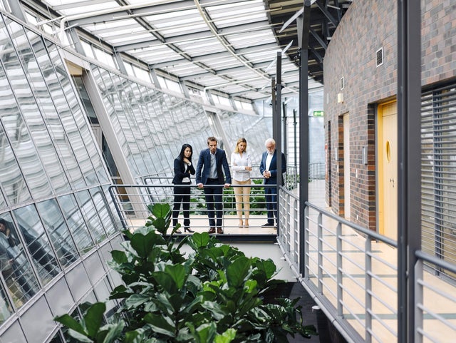 The picture shows four people in formal dress standing in a modern, light-flooded building. They are standing on a raised walkway with railings, surrounded by large glass windows. The windows extend along the entire length of the corridor and let in plenty of daylight.on the left of the picture is a woman with long, dark hair wearing a black trouser suit. She has her arms crossed in front of her chest and is looking down as if she is pensive. Next to her is a man in a dark blue suit, who is leaning on the railing with both hands and also looking down. Next to him is a woman with blonde hair in a white blazer and beige trousers. To her right is an older man with white hair and a beard, wearing a dark blue suit and interlocking fingers. The four people are discussing ista's building management system. Below the catwalk are large green plants that lend the room a natural element. On the right of the picture you can see a wall with dark red bricks and several doors. The whole space has a modern and open feel, with lots of natural light and a mix of industrial and natural elements.