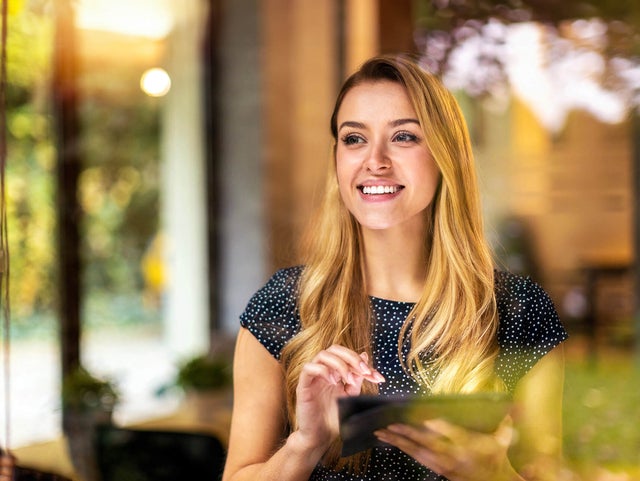 The picture shows a young woman looking through a window. She has long blonde hair and is wearing a dark blue, polka-dotted top. The woman is smiling and holding a tablet in both hands which is showing the Energy Audit Reporting from ista, her gaze directed slightly to the right. Green plants and a blurred light source can be seen in the background, giving the picture a warm and inviting atmosphere. The light source is located approximately in the centre of the background. The left background area shows a vertical black line, which is probably part of the window frame. The woman is standing relatively close to the window so that her reflection is partially visible in the glass. The foreground of the image is in focus while the background remains blurred, which draws the viewer's attention to the woman's smiling face and her tablet.