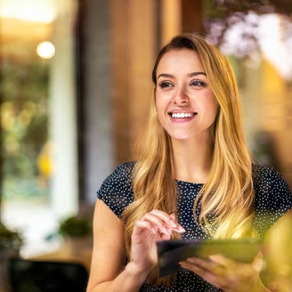 The picture shows a young woman looking through a window. She has long blonde hair and is wearing a dark blue, polka-dotted top. The woman is smiling and holding a tablet in both hands which is showing the Energy Audit Reporting from ista, her gaze directed slightly to the right. Green plants and a blurred light source can be seen in the background, giving the picture a warm and inviting atmosphere. The light source is located approximately in the centre of the background. The left background area shows a vertical black line, which is probably part of the window frame. The woman is standing relatively close to the window so that her reflection is partially visible in the glass. The foreground of the image is in focus while the background remains blurred, which draws the viewer's attention to the woman's smiling face and her tablet.