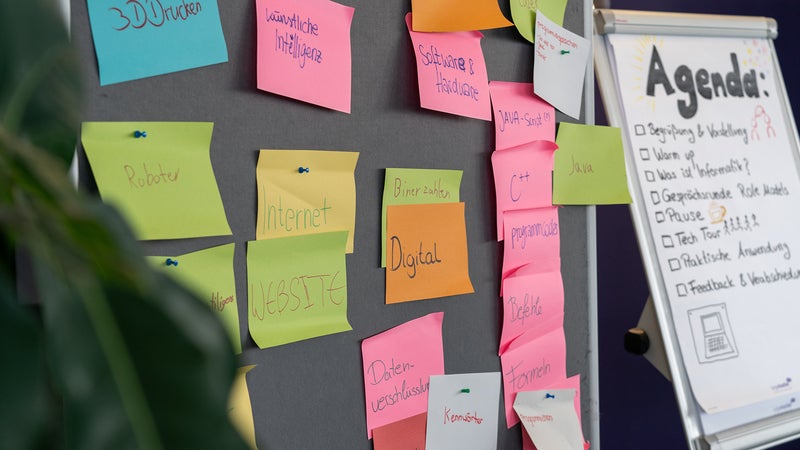 Eine Pinnwand, die mit bunten Haftnotizen beklebt ist, und ein Whiteboard mit einer auf Deutsch geschriebenen Agenda. Die Notizen enthalten Begriffe wie "3D-Drucken", "Künstliche Intelligenz", "Software & Hardware", "Binärzahlen", "Digital" und Programmiersprachen wie "Java", "C++" und "Python".