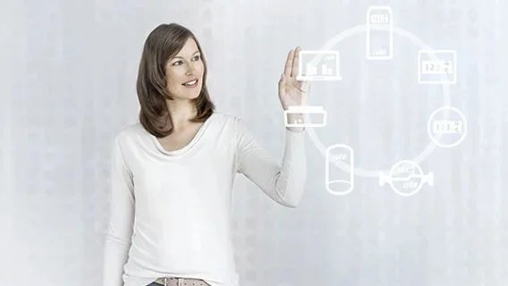 Eine Frau mit weißem Longsleeve betrachtet ein Hologramm welches einen Kreis mit mehreren verbundenen Geräten zeigt.