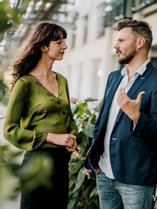 The picture shows a relaxed and friendly scene in which a woman and a man are chatting outdoors. The woman is on the left of the picture. She has long, slightly wavy dark hair and is wearing a green blouse with long sleeves and a V-neck. She holds her hands in front of her in a relaxed manner. The man on the right of the picture has short, dark hair. He is wearing a blue blazer over a white shirt and light-coloured jeans. As he speaks, he gestures with his right hand as if he is explaining or emphasising something. The background of the scene shows a modern building with large windows and light-coloured facades, suggesting an urban environment, possibly a terrace or courtyard. There are blurred green plants in the foreground and on the left-hand side of the picture, giving the image depth and creating a natural atmosphere. The light in the scene is natural and comes from the left side, which illuminates the scene well and creates a friendly, inviting mood. The overall atmosphere of the picture looks professional and at the same time relaxed, as if the two people are having a pleasant conversation. The combination of urban surroundings and nature conveys a sense of balance and harmony.