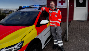 Der Mann, nun in einer roten Einsatzjacke des DRK, lehnt an einem DRK-Fahrzeug mit eingeschaltetem Blaulicht. Im Hintergrund ist ein Gebäude mit einem DRK-Logo zu sehen. Er lächelt in die Kamera und trägt Einsatzkleidung mit reflektierenden Streifen.