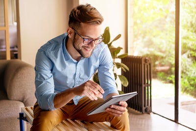 The picture shows a young man with glasses, leaning slightly forwards and sitting on a modern wooden bench. The bench is in a bright and cosy room. The man is wearing a light blue shirt and mustard-coloured trousers. He is holding a tablet in his hands and looking at ista's heat metering system. Behind the man, to the right of the picture, is a large floor-to-ceiling window that allows plenty of natural light into the room. A green garden with lush vegetation can be seen through the window. Next to the window is a large plant in a pot on the floor. To the right of the plant is a black radiator, which contrasts with the light and natural colours of the room. To the left of the man, part of a beige sofa is visible. Other parts of the room can be seen in the background on the left of the picture, possibly including a door or another window, which also bring light and space into the room.