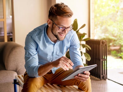 The picture shows a young man with glasses, leaning slightly forwards and sitting on a modern wooden bench. The bench is in a bright and cosy room. The man is wearing a light blue shirt and mustard-coloured trousers. He is holding a tablet in his hands and looking at ista's heat metering system. Behind the man, to the right of the picture, is a large floor-to-ceiling window that allows plenty of natural light into the room. A green garden with lush vegetation can be seen through the window. Next to the window is a large plant in a pot on the floor. To the right of the plant is a black radiator, which contrasts with the light and natural colours of the room. To the left of the man, part of a beige sofa is visible. Other parts of the room can be seen in the background on the left of the picture, possibly including a door or another window, which also bring light and space into the room.