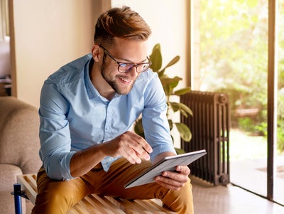 The picture shows a young man with glasses, leaning slightly forwards and sitting on a modern wooden bench. The bench is in a bright and cosy room. The man is wearing a light blue shirt and mustard-coloured trousers. He is holding a tablet in his hands and looking at ista's heat metering system. Behind the man, to the right of the picture, is a large floor-to-ceiling window that allows plenty of natural light into the room. A green garden with lush vegetation can be seen through the window. Next to the window is a large plant in a pot on the floor. To the right of the plant is a black radiator, which contrasts with the light and natural colours of the room. To the left of the man, part of a beige sofa is visible. Other parts of the room can be seen in the background on the left of the picture, possibly including a door or another window, which also bring light and space into the room.