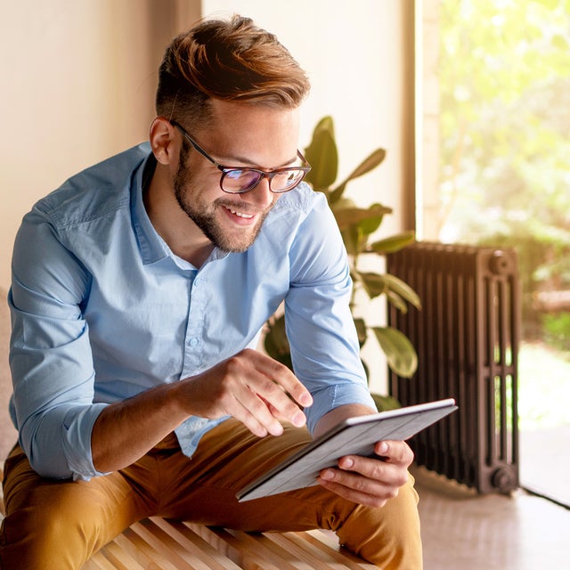 The picture shows a young man with glasses, leaning slightly forwards and sitting on a modern wooden bench. The bench is in a bright and cosy room. The man is wearing a light blue shirt and mustard-coloured trousers. He is holding a tablet in his hands and looking at ista's heat metering system. Behind the man, to the right of the picture, is a large floor-to-ceiling window that allows plenty of natural light into the room. A green garden with lush vegetation can be seen through the window. Next to the window is a large plant in a pot on the floor. To the right of the plant is a black radiator, which contrasts with the light and natural colours of the room. To the left of the man, part of a beige sofa is visible. Other parts of the room can be seen in the background on the left of the picture, possibly including a door or another window, which also bring light and space into the room.