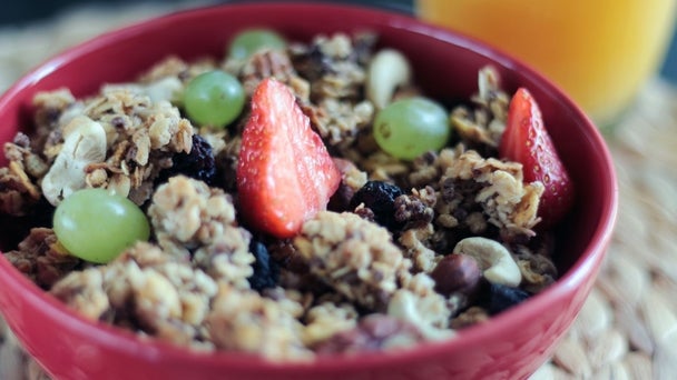 Eine Schale mit Müsli, Erdbeeren, Trauben und Nüssen steht auf einem Tisch, im Hintergrund ist ein Glas Orangensaft zu sehen.