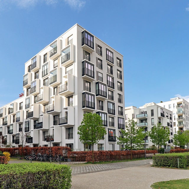 The picture shows a corner of a housing estate with white modern apartment buildings stretching from left to right across the picture. The buildings are characterised by an unusual architecture, as they alternately have protruding cubes with either large windows throughout the whole cube or set in balconies. The skyblue sky is filled with a few white clouds on the right side of the picture and in the foreground are red hedges and small trees that have been planted around the buildings, marking the property of the apartment buildings. There is a paved pedestrian path along the hedges and this is also bordered by another green hedge. The pedestrian path runs in a half circle in the bottom right of the picture, making a way through the grass that is planted there. 