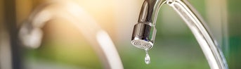 The picture shows a tap. The tap is made of shiny metal and has a curved shape that points downwards. At the end of the tap, where the water normally comes out, hangs a single drop of water that is about to fall. Another tap can be seen blurred in the background to the left of it. The entire background of the picture is blurred and shows a mixture of green and blue colours that could be reminiscent of an outdoor area or a garden. The light in the picture is warm and appears to be coming from the top left, casting a soft, golden glow on the tap. This creates a feeling of freshness and clarity, as if it is a sunny morning. In the foreground of the image, below the tap, there is a blurred area which may represent a sink or basin.