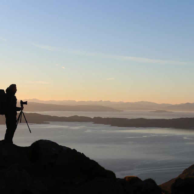Silhouette eines Fotografen mit Rucksack und Stativ auf einem Hügel bei Sonnenuntergang, mit Blick über eine Küstenlandschaft und Berge im Hintergrund.