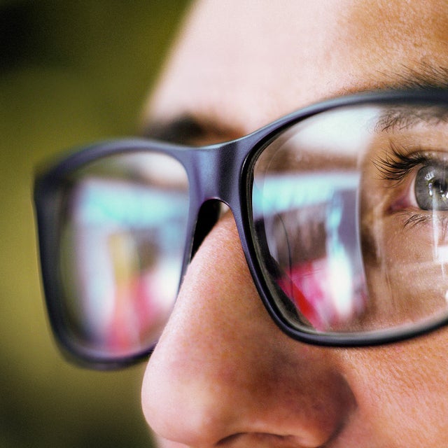 The picture shows a close-up of the eye area of a person wearing square, black glasses. The person is looking from the right side of the picture to the left. You can only see the person's left eye, which is grey-blue and surrounded by dark eyelashes. The focus of the picture is on the recognisable eye, the rest of the picture is blurry.