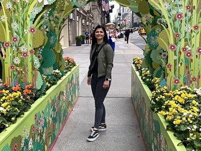 Eine Frau steht unter einem dekorativen, blumenverzierten Torbogen auf einer belebten Straße in einer Stadt. Um sie herum blühen bunte Blumen in Hochbeeten, und im Hintergrund sind elegante Gebäude und die amerikanische Flagge zu sehen.