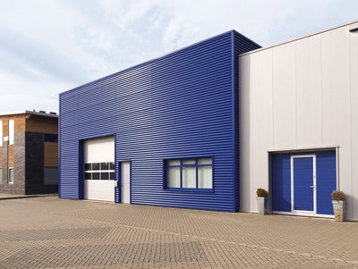 The picture shows a central warehouse with a horizontally fluted blue steel façade and a large white garage entrance for lorries on the left-hand side of the building. This entrance is decorated with eight windows. Next to the garage door is a simple white entrance door. On the right-hand side of the building, a window is divided into three sections and covered with white shutters on the inside. Attached to the warehouse on the right is a slightly lower, white fluted building, which also has a steel façade. The extension has a garage door at car height, which also contains a built-in entrance door. It is the same blue colour as the warehouse façade. To the left and right of this garage door are two taller pots with spherical plants. A drainage pipe runs along the white extension on the right-hand edge of the picture. In the left quarter of the picture, next to the blue warehouse, a modern house can be seen. It has an asymmetrical wood and stone façade decorated with windows of different sizes. In the lower third of the picture there is a kind of driveway and car park for the warehouse. The sky is bluish and slightly cloudy.