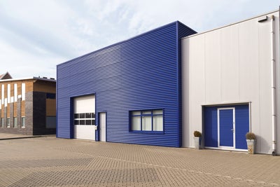 The picture shows a central warehouse with a horizontally fluted blue steel façade and a large white garage entrance for lorries on the left-hand side of the building. This entrance is decorated with eight windows. Next to the garage door is a simple white entrance door. On the right-hand side of the building, a window is divided into three sections and covered with white shutters on the inside. Attached to the warehouse on the right is a slightly lower, white fluted building, which also has a steel façade. The extension has a garage door at car height, which also contains a built-in entrance door. It is the same blue colour as the warehouse façade. To the left and right of this garage door are two taller pots with spherical plants. A drainage pipe runs along the white extension on the right-hand edge of the picture. In the left quarter of the picture, next to the blue warehouse, a modern house can be seen. It has an asymmetrical wood and stone façade decorated with windows of different sizes. In the lower third of the picture there is a kind of driveway and car park for the warehouse. The sky is bluish and slightly cloudy.