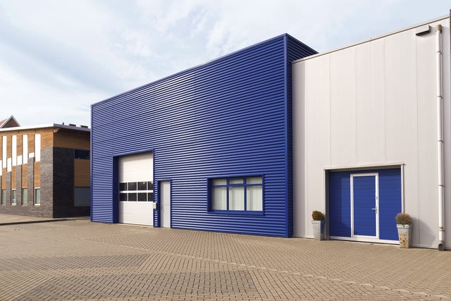The picture shows a central warehouse with a horizontally fluted blue steel façade and a large white garage entrance for lorries on the left-hand side of the building. This entrance is decorated with eight windows. Next to the garage door is a simple white entrance door. On the right-hand side of the building, a window is divided into three sections and covered with white shutters on the inside. Attached to the warehouse on the right is a slightly lower, white fluted building, which also has a steel façade. The extension has a garage door at car height, which also contains a built-in entrance door. It is the same blue colour as the warehouse façade. To the left and right of this garage door are two taller pots with spherical plants. A drainage pipe runs along the white extension on the right-hand edge of the picture. In the left quarter of the picture, next to the blue warehouse, a modern house can be seen. It has an asymmetrical wood and stone façade decorated with windows of different sizes. In the lower third of the picture there is a kind of driveway and car park for the warehouse. The sky is bluish and slightly cloudy.