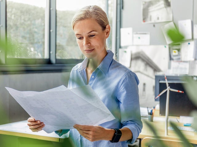 The picture shows a room, with windows in the top left side. On the top right side are many papers pasted on the wall and below them are two office desks. The focus is on a woman with a blue shirt, standing in the middle of the picture, right before the desks, holding a few sheets of the energy efficiency certificates.
