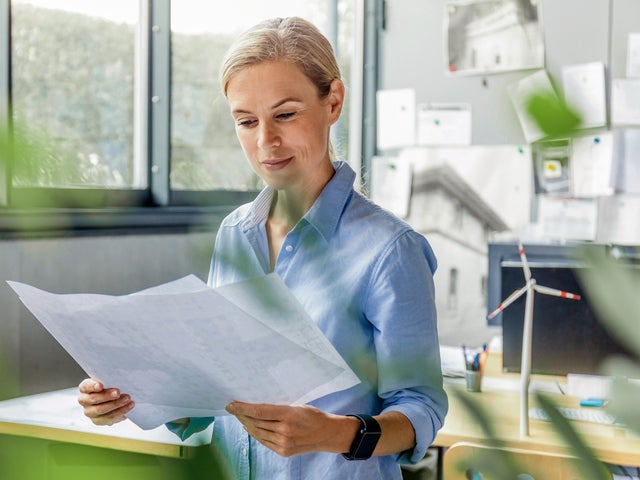 The picture shows a room, with windows in the top left side. On the top right side are many papers pasted on the wall and below them are two office desks. The focus is on a woman with a blue shirt, standing in the middle of the picture, right before the desks, holding a few sheets of the energy efficiency certificates.