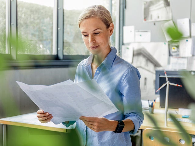 The picture shows a room, with windows in the top left side. On the top right side are many papers pasted on the wall and below them are two office desks. The focus is on a woman with a blue shirt, standing in the middle of the picture, right before the desks, holding a few sheets of the energy efficiency certificates.