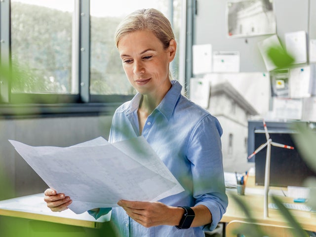 The picture shows a room, with windows in the top left side. On the top right side are many papers pasted on the wall and below them are two office desks. The focus is on a woman with a blue shirt, standing in the middle of the picture, right before the desks, holding a few sheets of the energy efficiency certificates.