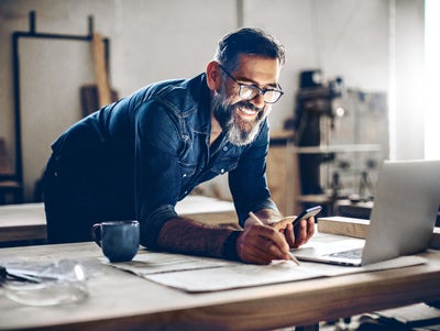The picture shows a middle-aged man working in a workshop or studio. He has dark hair, wears glasses and his face is framed by a full, well-groomed grey beard. He wears a dark blue denim shirt and smiles broadly.The man is leaning over a large work table while holding a smartphone in his left hand looking at ista’s operating cost billing and making notes on the paper with his right hand. There is an open laptop on the table in front of him. Next to the laptop is a blue coffee cup.The background of the workshop is blurred, but you can recognise shelves and tools that indicate a manual or creative activity. The light streaming in from the right through a window illuminates the scene and creates a warm, inviting atmosphere.The man appears focussed and content, which is conveyed by his smile and relaxed posture.
