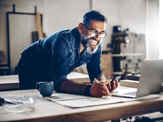 The picture shows a middle-aged man working in a workshop or studio. He has dark hair, wears glasses and his face is framed by a full, well-groomed grey beard. He wears a dark blue denim shirt and smiles broadly.The man is leaning over a large work table while holding a smartphone in his left hand looking at ista’s operating cost billing and making notes on the paper with his right hand. There is an open laptop on the table in front of him. Next to the laptop is a blue coffee cup.The background of the workshop is blurred, but you can recognise shelves and tools that indicate a manual or creative activity. The light streaming in from the right through a window illuminates the scene and creates a warm, inviting atmosphere.The man appears focussed and content, which is conveyed by his smile and relaxed posture.