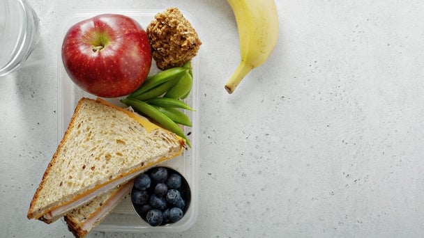 Eine Lunchbox mit belegtem Sandwich, Apfel, Banane, Blaubeeren und Gemüsesticks liegt auf einer hellen Tischplatte.