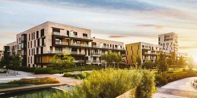 The picture shows a residential complex in the centre, stretching from the left to the right edge of the picture. The various buildings look very similar on the outside and have a white-orange brick façade. The individual flats have balconies with glass railings. The balconies are separated from each other by frosted glass walls and are furnished differently, giving each flat an individual touch. Most of the visible buildings have four floors, but vary in width so that different numbers of flats are visible on each floor. In the right quarter of the picture, in contrast to the others, another building rises up, which has the same style as the other buildings in the residential complex. A park can be seen in front of the residential complex in the lower half of the picture. Grasses and bushes surround the residential complex, accompanied by a pavement adorned with scattered trees. There are also benches and lanterns on the pavement. An artificial pond can be seen in the bottom left-hand corner, surrounded by a wooden walkway with glass railings. The sunset can be seen on the right-hand edge of the picture, giving the sky a slight colour gradient, from a reddish tone on the right to a bluish tone on the left.