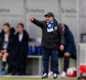 Der Mann steht am Spielfeldrand eines Fußballplatzes, trägt Sportkleidung mit Vereinslogo und einen blauen Fanschal. Er zeigt mit ausgestrecktem Arm in eine Richtung, während Spielerinnen auf der Bank im Hintergrund sitzen.
