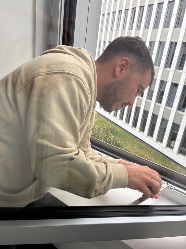 Ein Mann beugt sich an einem geöffneten Fenster nach vorne und führt eine handwerkliche Arbeit aus.