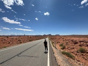Eine Frau steht auf einer langen, geraden Straße in einer weiten Wüstenlandschaft unter einem strahlend blauen Himmel. Im Hintergrund erstrecken sich Felsenformationen – der berühmte Forrest-Gump-Point in Monument Valley, USA.