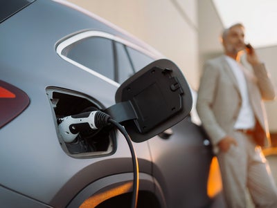 The picture shows an electric car that is being charged. In the foreground is the car's charging socket, into which a charging cable is plugged. The cable is connected to an ista EV charging station, which is outside the picture. In the background, slightly out of focus, is a man in a light-coloured suit talking on the phone. He has a relaxed posture and is leaning slightly against the car. The scene probably takes place in an urban environment or in a company car park, which is indicated by the modern buildings in the background. The image conveys an atmosphere of modern technology and environmentally friendly mobility.