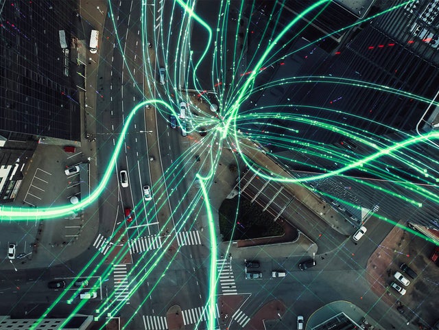 The picture shows a bird's eye view of an urban intersection. There are several wide streets that meet in the centre of the picture and form a complex network of traffic routes. Numerous curved, bright blue lines run across these streets, symbolising the flow of data or information exchange. These lines start from different points and concentrate in the centre of the intersection, creating a radial pattern. At the bottom of the picture, several moving cars can be seen travelling along the road. On the left-hand side of the picture is a building with a car park where several cars are parked. On the right-hand side of the picture, tall, modern office buildings with large window fronts are visible, emphasising the urban environment. The blue lines that dominate the image convey a dynamic sense of connectivity and technology, while the underlying urban infrastructure provides the physical basis for this digital exchange.