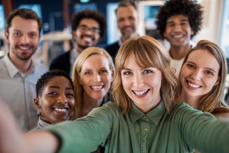 The image shows a group of diverse people smiling and taking a group selfie. They are standing close together in an indoor setting that appears to be an office or a collaborative workspace. The people in the photo represent a variety of ages, genders, and ethnic backgrounds, reflecting a diverse and inclusive environment. The atmosphere is cheerful and positive, suggesting a sense of camaraderie and teamwork among the group members.