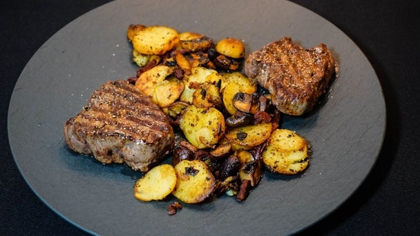 Ein Teller mit zwei gegrillten Steaks, Bratkartoffeln und Pilzen auf dunklem Untergrund.