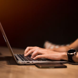 A person types on a laptop in a dimly lit environment, with a smartwatch on their wrist and a smartphone placed on the table beside them. The warm, focused lighting emphasizes productivity and technology use, highlighting a modern workspace or remote work setting. The image conveys themes of digital connectivity, work-life balance, and the integration of smart devices in daily tasks.